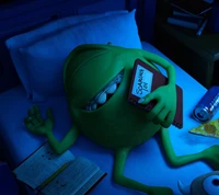 A green, round monster lounging on a bed, holding a game controller, with a playful expression in a dimly lit room.