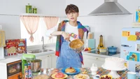 Huening Kai from Tomorrow X Together joyfully prepares breakfast in a colorful kitchen filled with delicious treats.