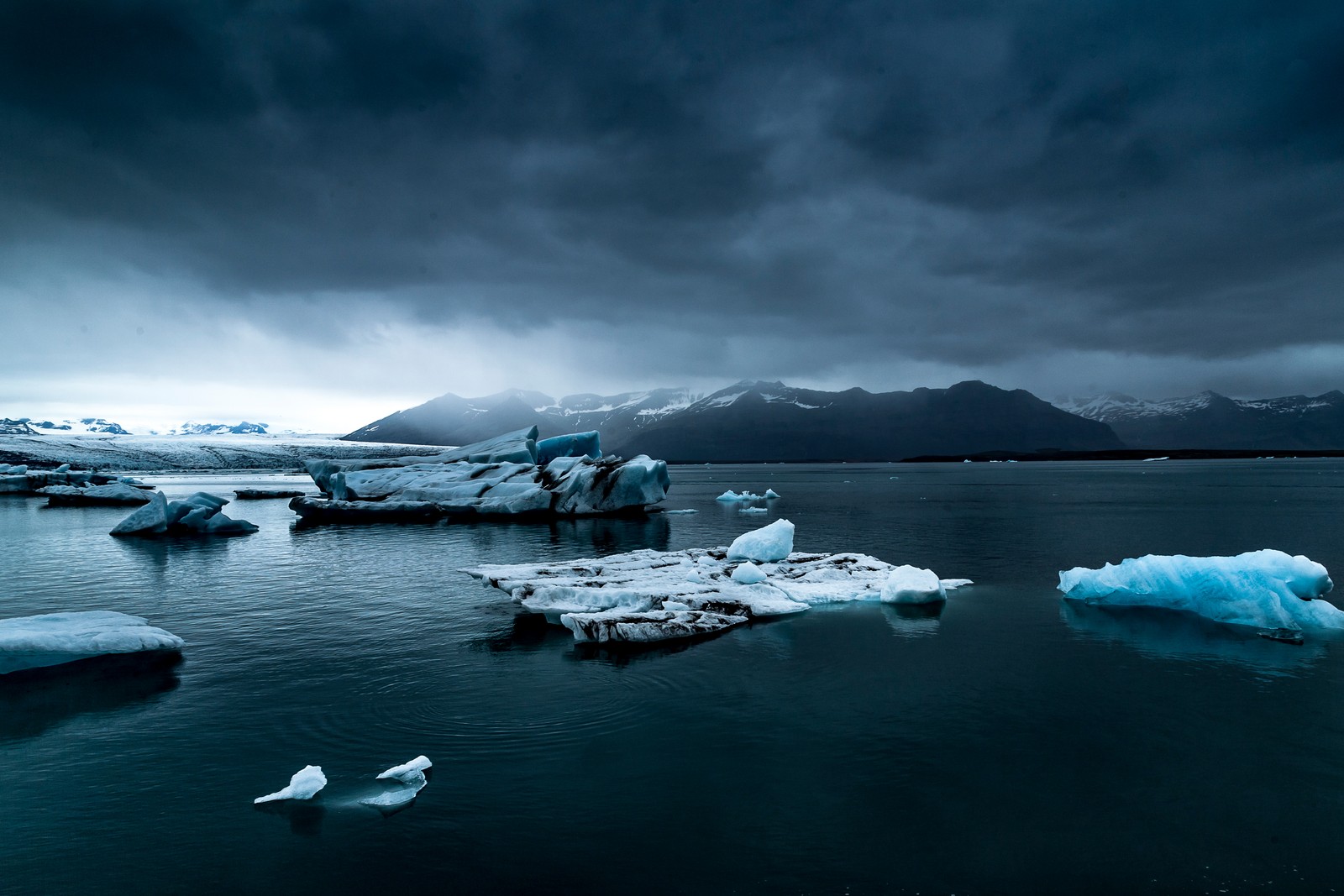 Vista aérea de icebergs flotando en un lago con montañas al fondo (lago glaciar, lago congelado, invierno, cielo nublado, frío)