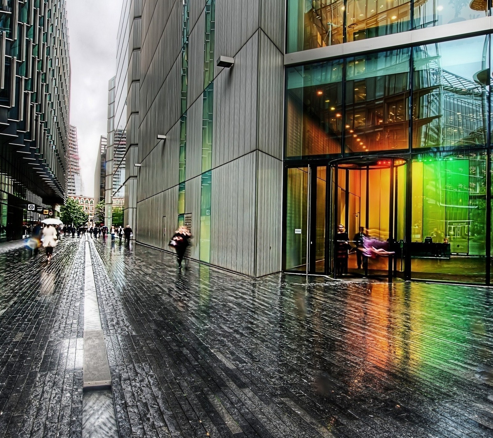 Des gens marchant sur un trottoir mouillé devant un bâtiment. (abstrait, génial, beauté, ville, coloré)