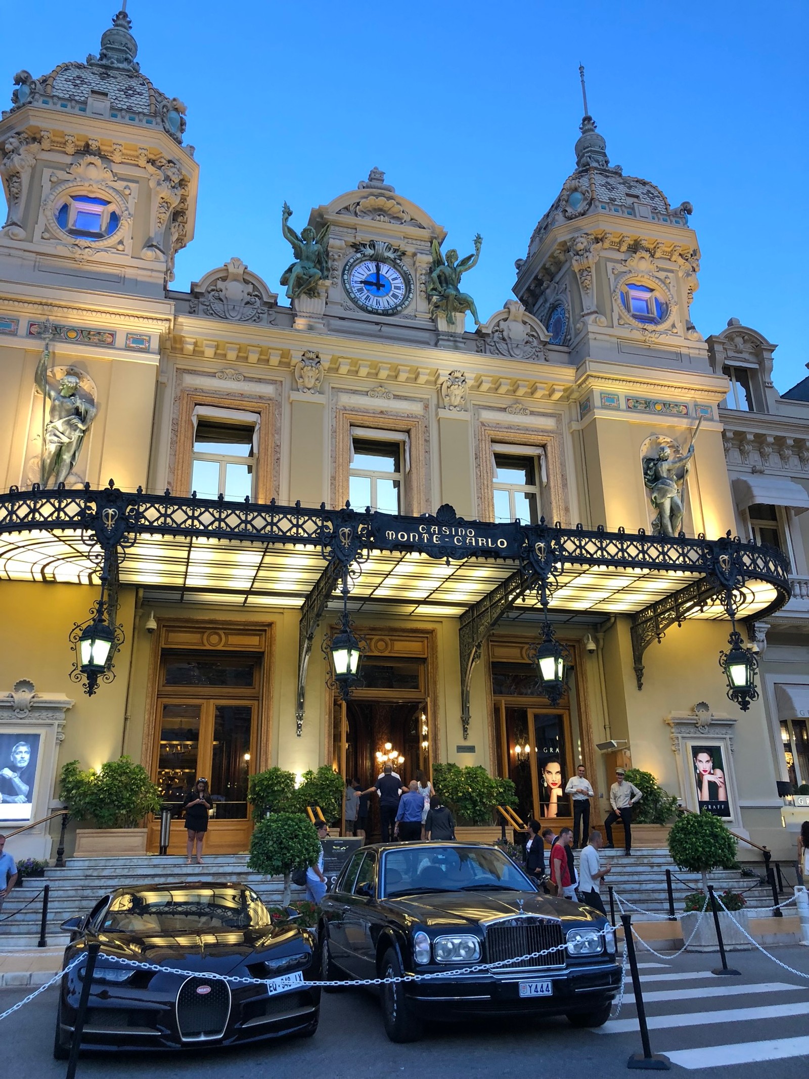 Автомобили припаркованы у большого здания с часами на крыше. (monte carlo casino, решетка, бампер, дверь, капюшон)