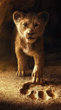 A young lion cub stands poised in a cave, gazing intently at a large paw print in the sandy ground, symbolizing its connection to legacy and future.