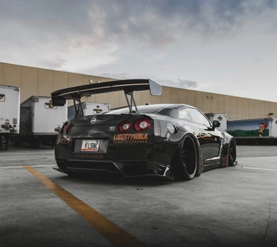 Nissan GT-R modificado com um kit de carroceria larga e um grande spoiler traseiro, estacionado contra um fundo industrial.