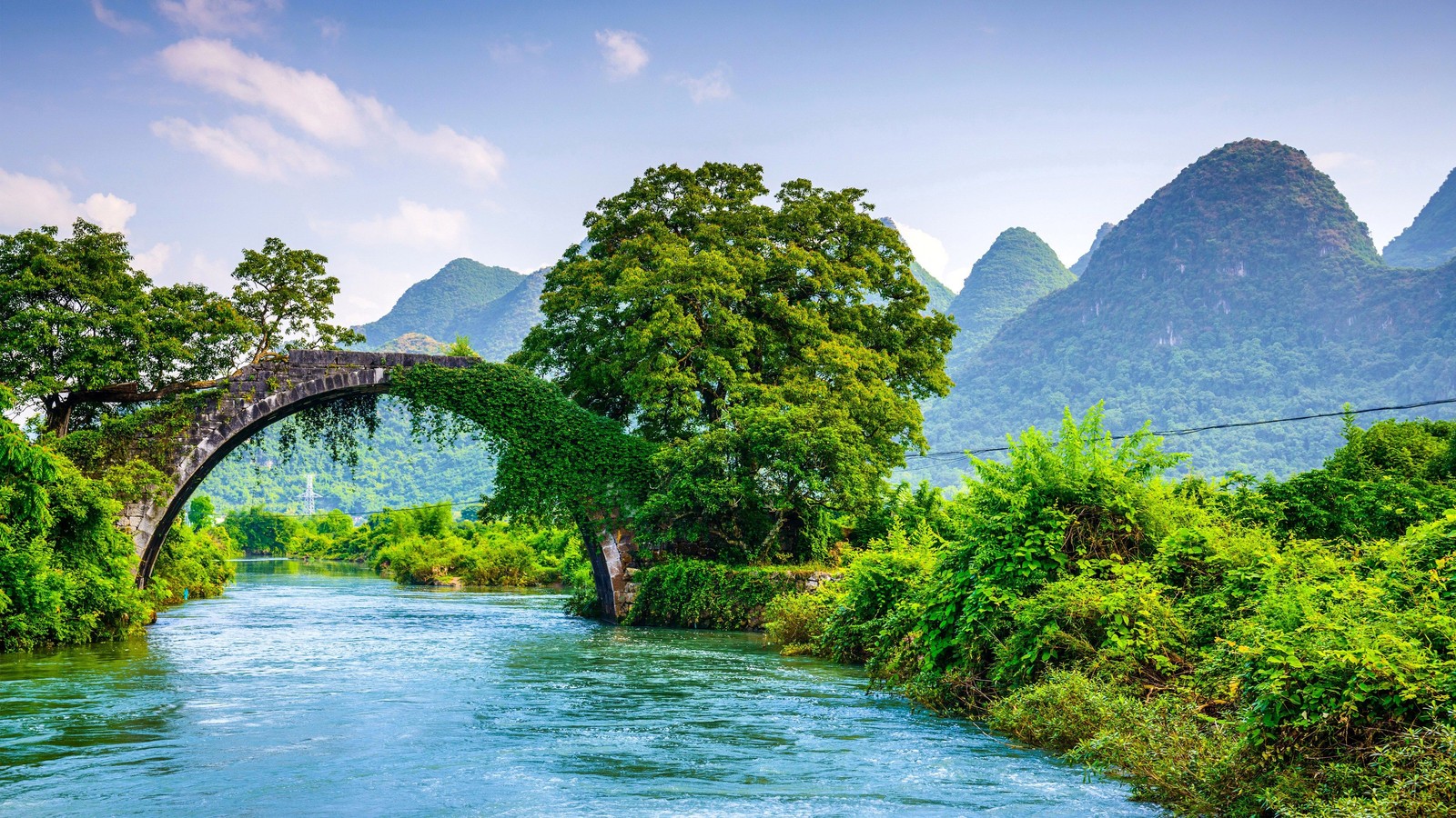 A bridge over a river surrounded by lush green trees and mountains (natural landscape, arch bridge, nature, river, water)