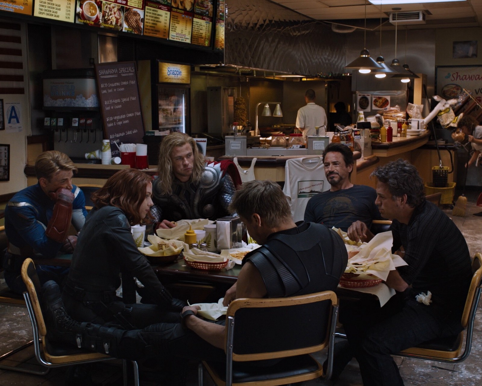 Several people sitting at a table eating food in a restaurant (assemble, avengers, captain, green arrow, hulk)