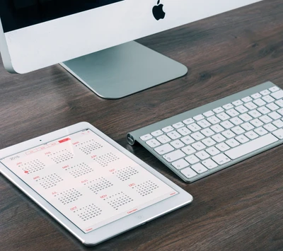 Apple Mac-Setup mit einem Kalender auf einem iPad und einer eleganten Tastatur auf einem Holztisch.