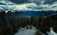 Dramatic mountain landscape with snow-capped peaks, dense evergreen trees, and a cloudy sky over a winding valley in Banff National Park.