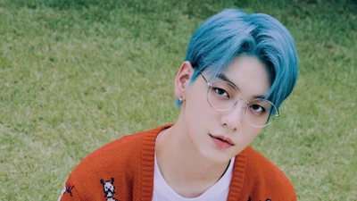 Soobin do TXT em uma sessão de fotos vibrante, apresentando seu cabelo azul marcante e óculos estilosos contra um fundo verde exuberante.