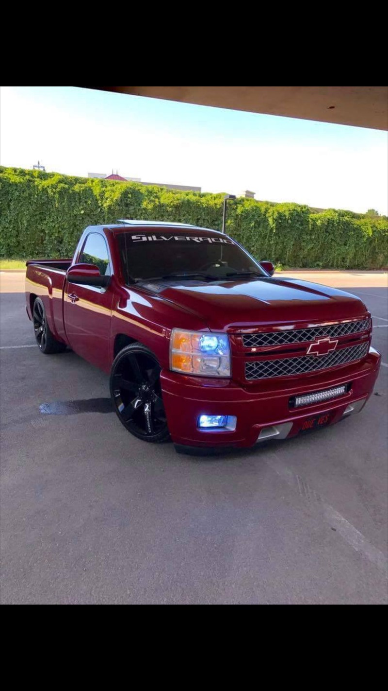 Un camión rojo estacionado en un aparcamiento con un seto verde (silverado, cabina simple)