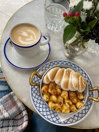 Cappuccino servido con una selección de pasteles dulces en un plato decorativo, acompañado de un arreglo floral y un vaso de agua, colocado sobre una mesa de mármol.