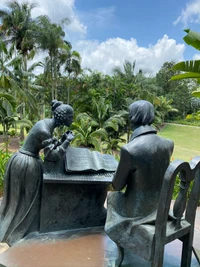 Escultura de bronce de una mujer y un hombre leyendo juntos en un jardín exuberante