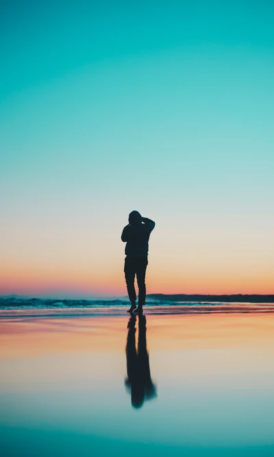 Silhueta de uma pessoa em pé em uma praia durante o pôr do sol, refletindo cores vibrantes de azul e laranja na água.