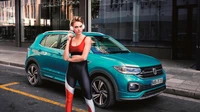 Cara Delevingne poses confidently beside a vibrant turquoise Volkswagen T-Cross, showcasing the compact SUV's sleek design against an urban backdrop.