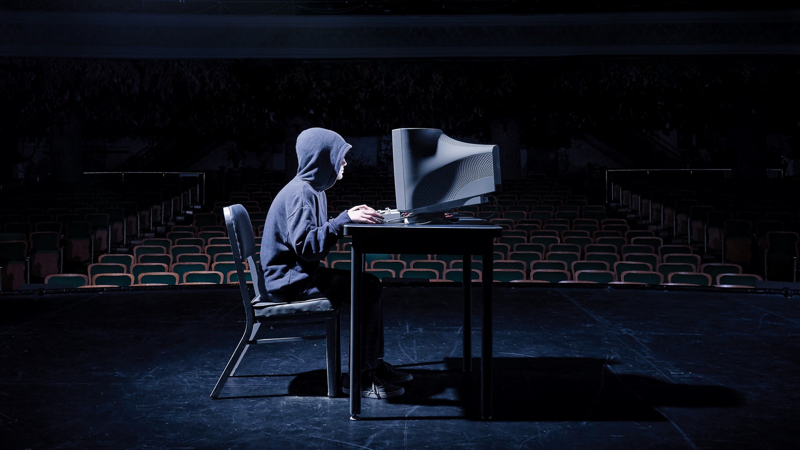 Arafed man in hoodie sitting at desk with computer in dark auditorium (hacker, black, light, darkness, stage)