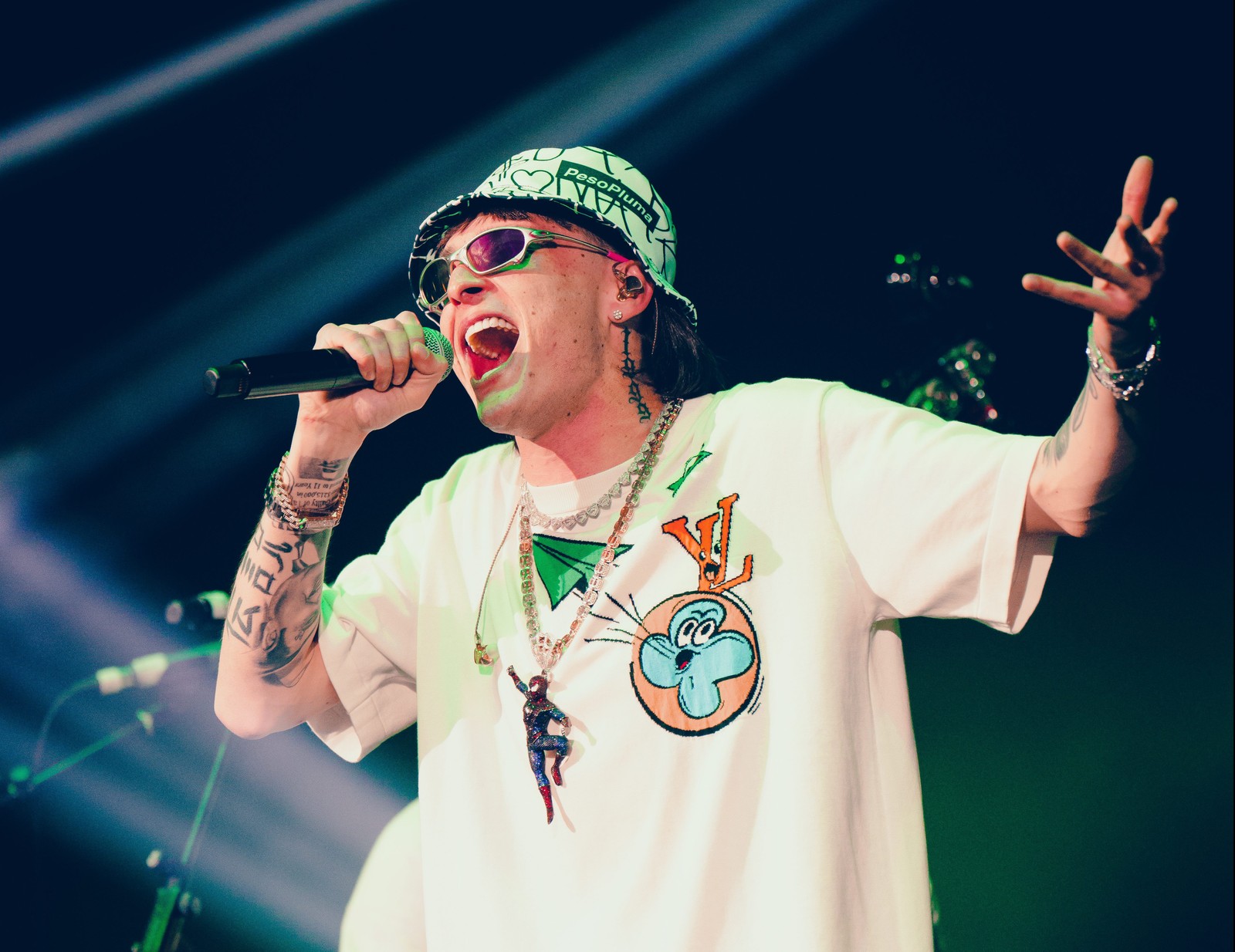 Un hombre borroso con un sombrero verde y gafas de sol cantando en un micrófono (peso pluma, cantante mexicano, rapero, concierto en vivo, música)