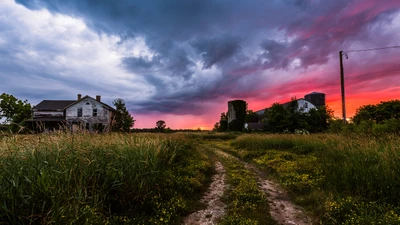 Coucher de soleil rural dans le Wisconsin illuminant un champ herbeux avec une ferme abandonnée et des silos contre un ciel dramatique.