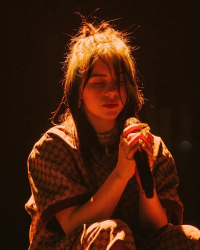 Billie Eilish en una pose contemplativa, iluminada por luces cálidas del escenario, vistiendo un atuendo estampado.