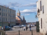 Medieval architecture in a vibrant town square, featuring a pedestrian-friendly street lined with cars, historic buildings, and a prominent steeple against a blue sky.