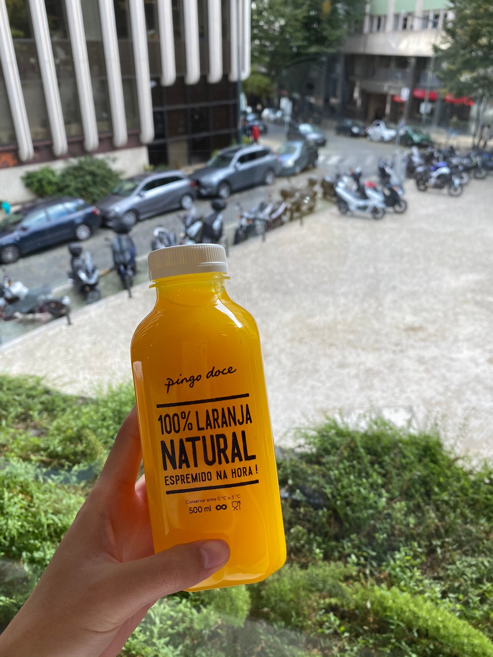 Alguien sostiene una botella de jugo de naranja frente a un edificio (botella, rey, fluido, líquido, pieza de automóvil)