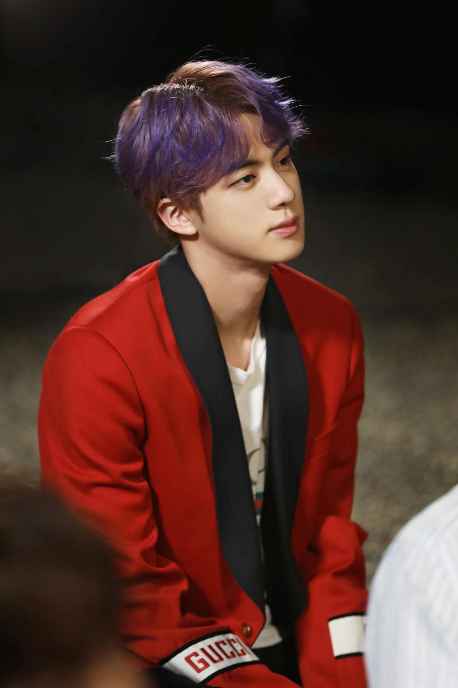 Un homme arabe en veste rouge et cravate noire assis sur un banc (zip militaire, bts, jin)