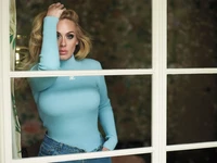 Adele olha pensativamente através de uma janela, exibindo seu estilo característico com uma blusa azul clara e jeans casuais, emoldurada por uma suave luz natural.