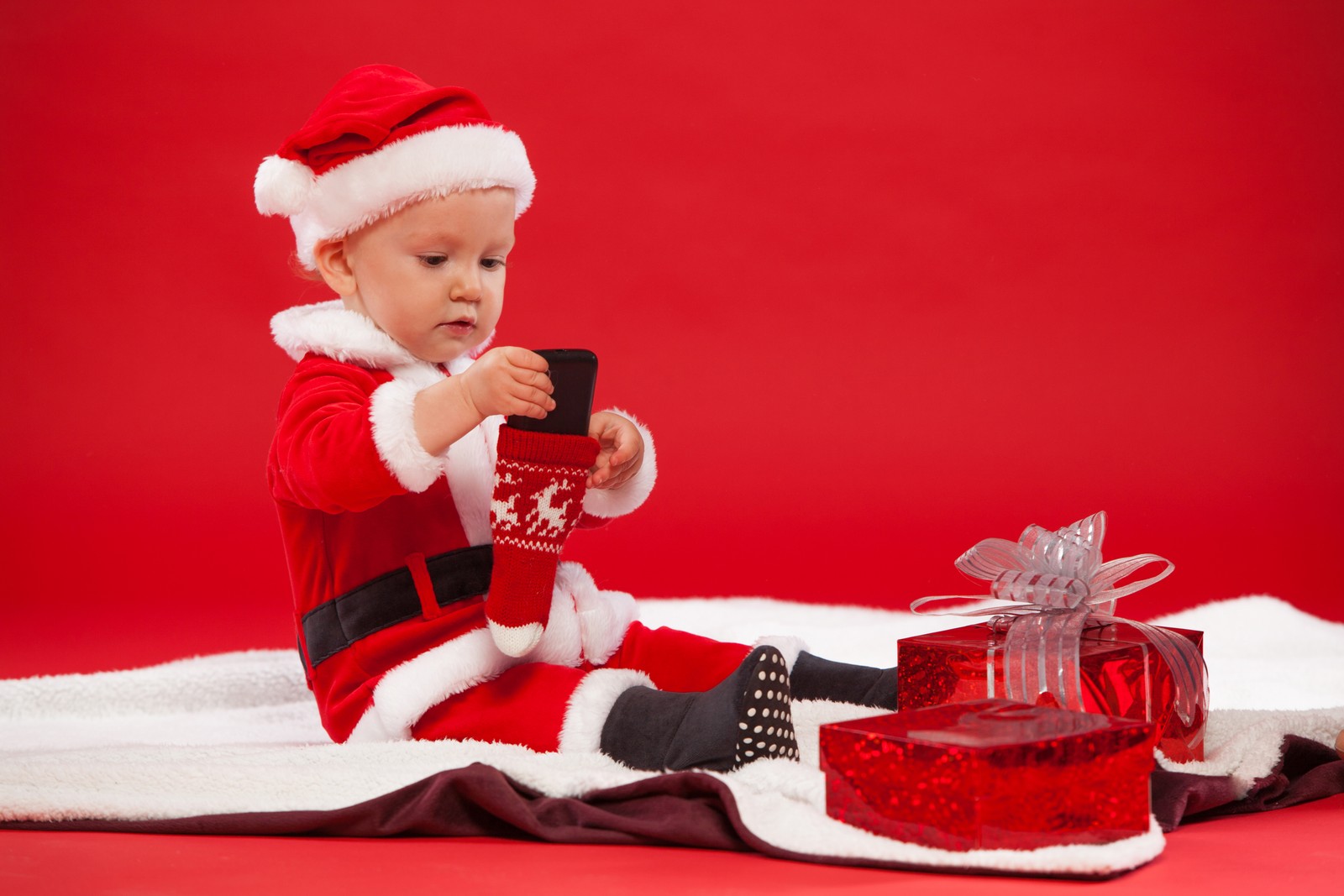 Un bébé en costume de père noël assis sur une couverture avec un téléphone portable (noël, père noël, cadeau, vacances, enfant)