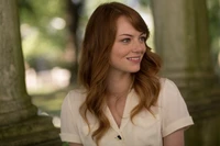 Emma Stone with a warm smile, showcasing her long brown hair and beauty in a serene outdoor setting, captured in a film still from 2015.