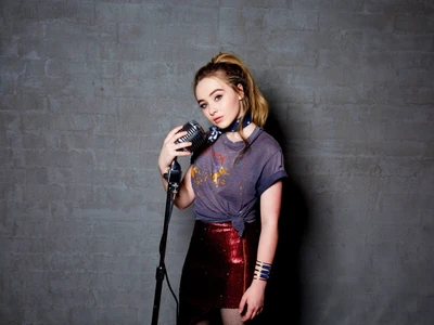 Sabrina Carpenter posing with a vintage microphone, showcasing a stylish look in a sparkly skirt and casual top against a textured gray background.