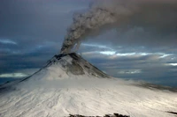 Stratovolcan enneigé avec un panache de cendres et de fumée s'élevant dans un ciel nuageux, montrant une activité volcanique au milieu d'un paysage de toundra austère.