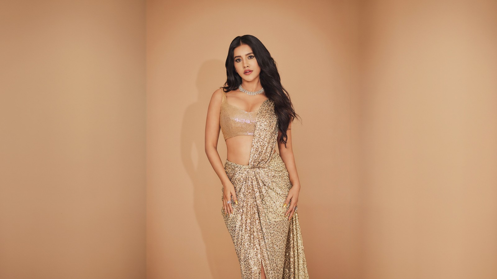 Eine frau in einem goldenen kleid, die vor einer beigen wand steht (nabha natesh, sari, saree, indische schauspielerin, goldener hintergrund)