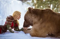 Uma mulher em cosplay interage gentilmente com um urso pardo em uma paisagem nevada.