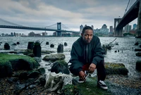 Kendrick Lamar poses contemplatively by the water's edge, with the iconic Brooklyn Bridge and a city skyline in the background, capturing a moment of artistry and urban life.