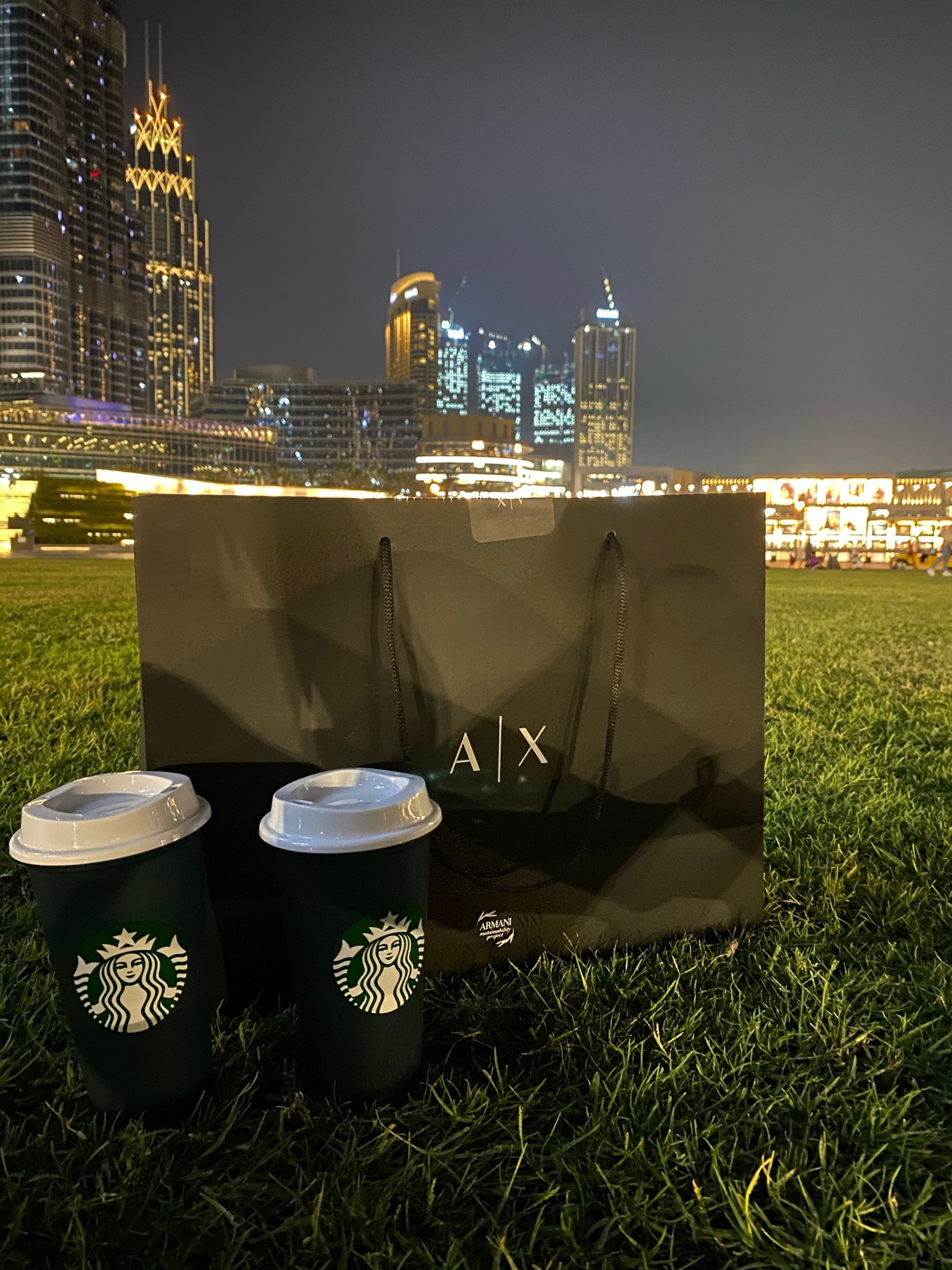 Taza de starbucks y bolsa de papel sentadas en el césped frente a un horizonte de ciudad (starbucks, bloque de torre, edificio, torre, noche)