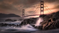 Puente Golden Gate al anochecer: olas rompen en la costa rocosa bajo un cielo dramático