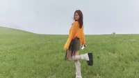 Ningning from aespa striking a playful pose in a vibrant orange sweater and chic knee-high boots against a lush green backdrop.