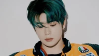 Jaehyun von NCT 127 mit auffälligem grünem Haar, das eine coole und nachdenkliche Ausstrahlung in einer lebhaften orangefarbenen Jacke ausstrahlt.