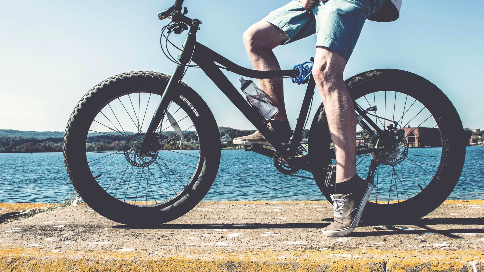 Homem árabe andando de bicicleta em um píer perto da água. (bicicleta, selim de bicicleta, roda de bicicleta, ciclismo, quadro de bicicleta)