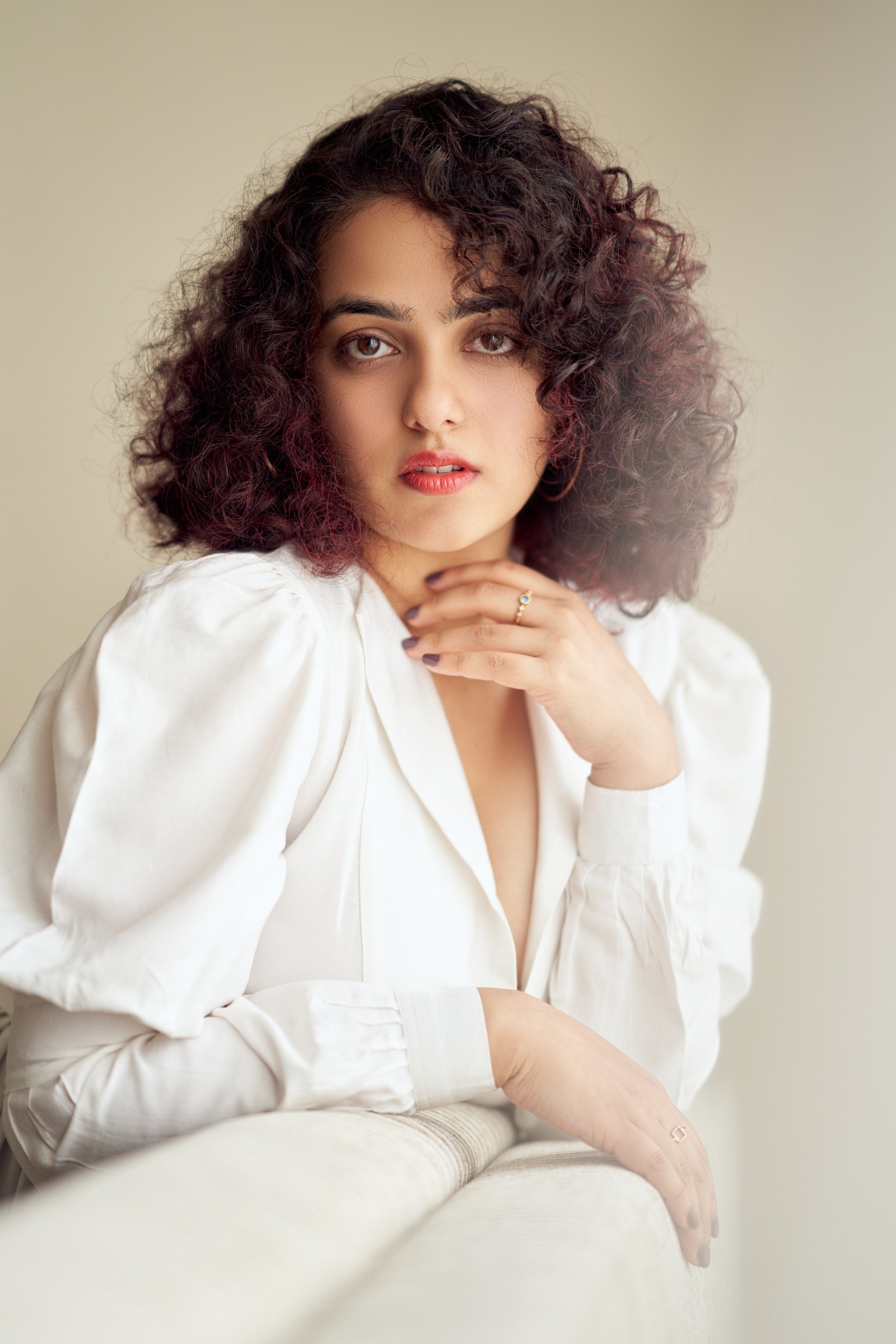 Une femme arabe avec des cheveux bouclés assise sur un canapé avec la main sur la poitrine (nithya menen, actrice indienne, portrait, belle actrice, 5k)