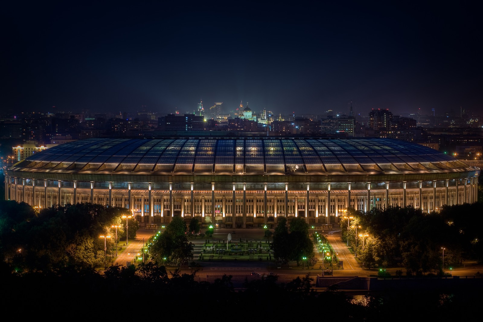 Uma vista de um estádio iluminado à noite com uma cidade ao fundo. (estádio, copa do mundo 2018, 2018 world cup, local desportivo, noite)