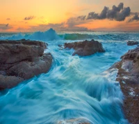 Dynamische Meereslandschaft bei Sonnenuntergang mit brechenden Wellen gegen die felsige Küste, die die lebendigen Farben des Himmels und des Ozeans einfängt.