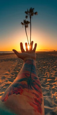 Mão alcançando um pôr do sol sobre a praia com tatuagens vibrantes e palmeiras em silhueta