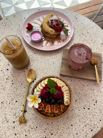 Petit-déjeuner coloré avec un bol de smoothie vibrant orné de fruits frais et du mot "BALI", accompagné d'un dessert empilé et d'une boisson rafraîchissante, le tout magnifiquement présenté sur de la vaisselle élégante.