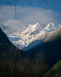 Schneebedeckte Gipfel erheben sich majestätisch über üppige Täler, umrahmt von einem klaren blauen Himmel und sanften Wolken, die die atemberaubende Schönheit einer Berglandschaft zeigen.