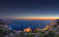 Una vista aérea impresionante del Salón Náutico de Mónaco al anochecer, que muestra las vibrantes luces de la ciudad a lo largo de la costa, un sereno paisaje marino y un impresionante horizonte bañado en los cálidos tonos del atardecer.