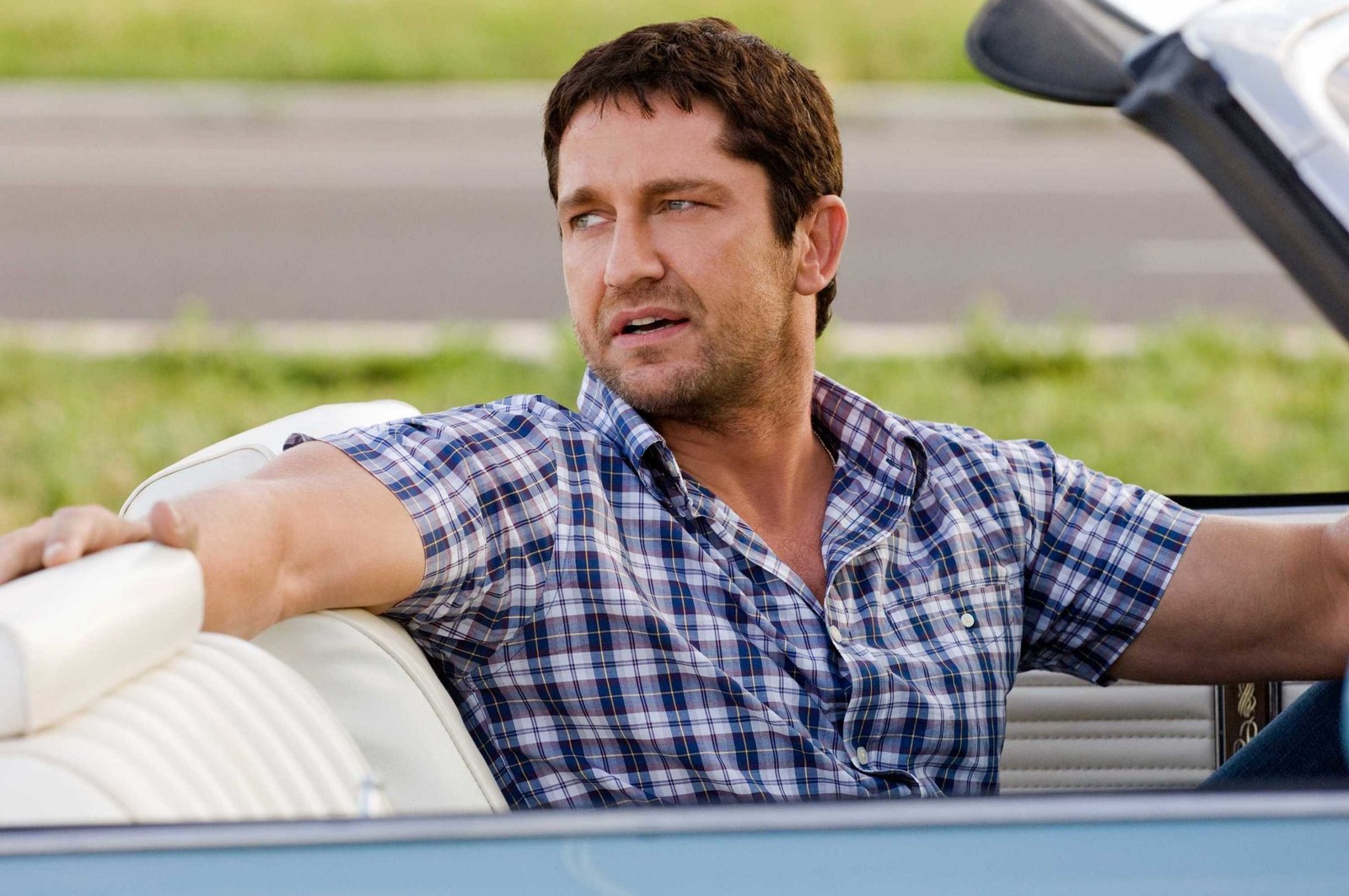 Un hombre árabe con una camisa azul sentado en un coche azul (gerard butler, músculo, sentado, coche, hollywood)