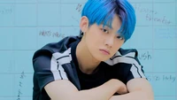 Yeonjun von TXT mit blauen Haaren, der eine fesselnde Pose vor einem pastellfarbenen Hintergrund zeigt.