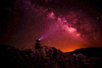 A silhouette gazes at the vibrant Milky Way, illuminating the night sky with cosmic wonders amidst a backdrop of darkness and distant galaxies.