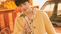 Woozi from Seventeen in a cheerful yellow coat, holding a rose, radiating warmth and charm.