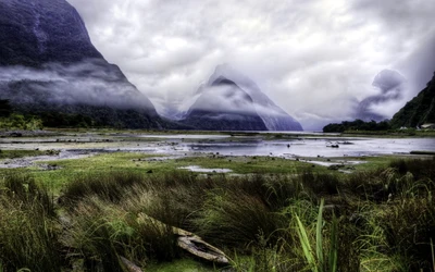 Fjord majestuoso de alta montaña con montañas brumosas reflejándose en las aguas tranquilas, rodeado de vegetación exuberante y un cielo dramático.