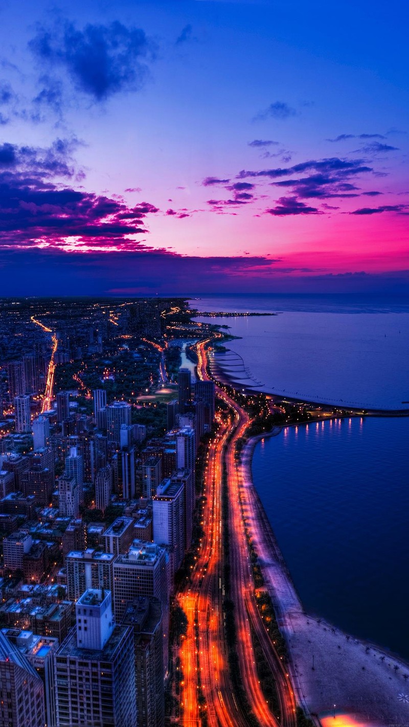 Vista aérea de una ciudad al atardecer con una playa y un cuerpo de agua. (playa, luces, noche, camino)